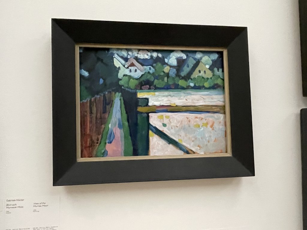 Bild von Gabriele Münter. Das Bild ist auch in Tupftechnik gestaltet, es ist eine Art Weide in hellrosa, gelben oder orangen Tönen getupft. Die Weide ist von einem Zaun umgeben, dann kommt ein Weg und dann wieder ein brauner Zaun.