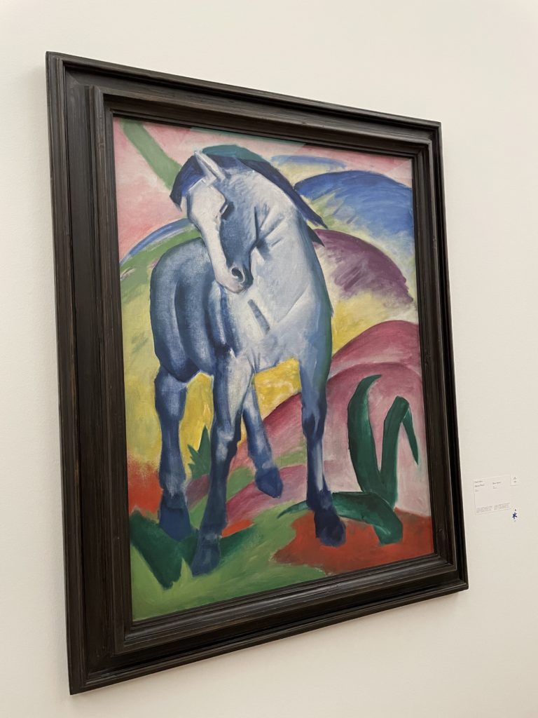 Ein Bild von Franz Marc. Ein blaues Pferd ist darauf zu sehen, dazu Hügel in Farben wie blau, grün, lila, gelb und rot. Dazu blättrige Pflanzen in Grün.